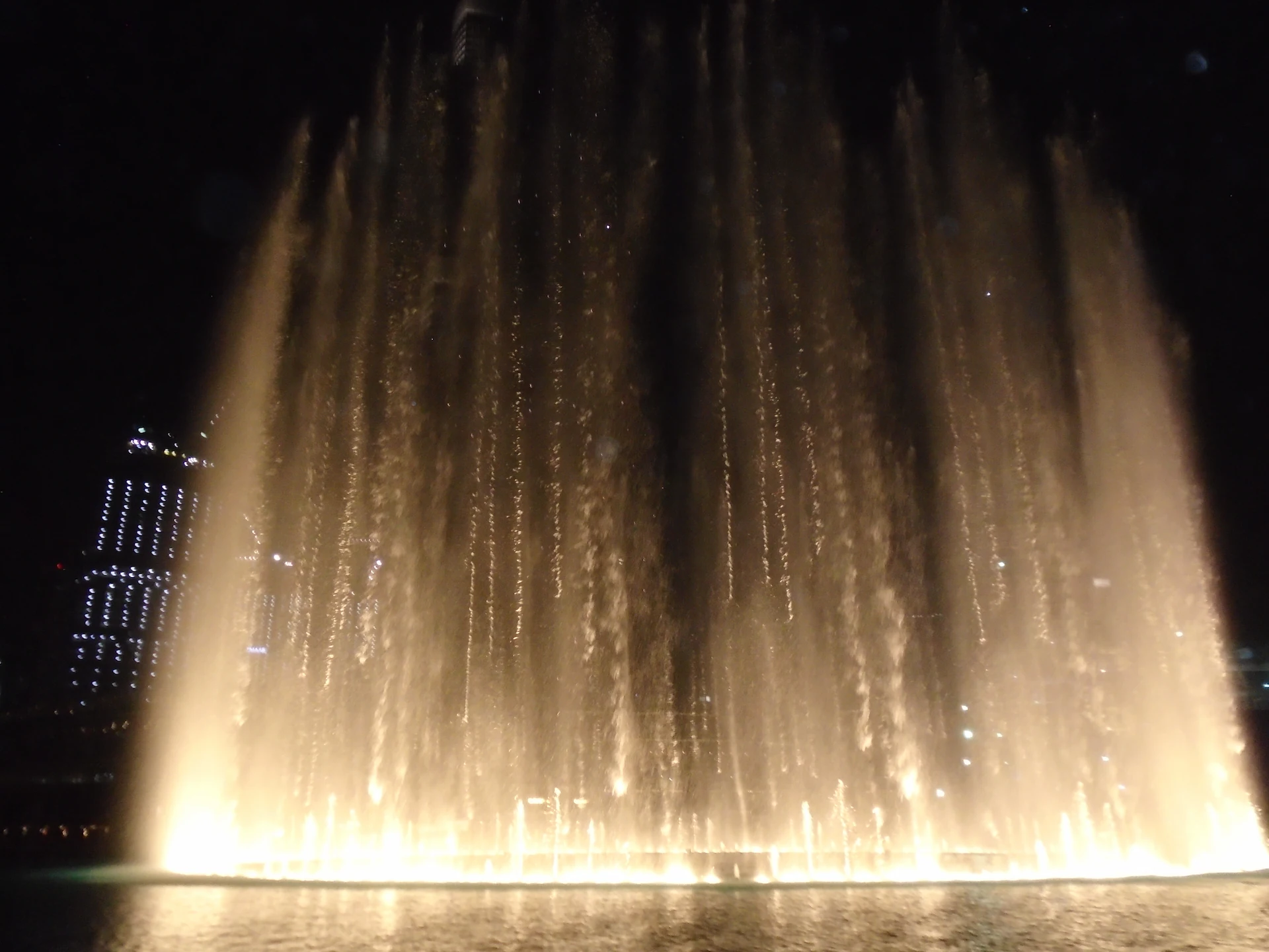 Dubai Fountain at night with water jets illuminated by lights performing over a large pool with city skyline in background