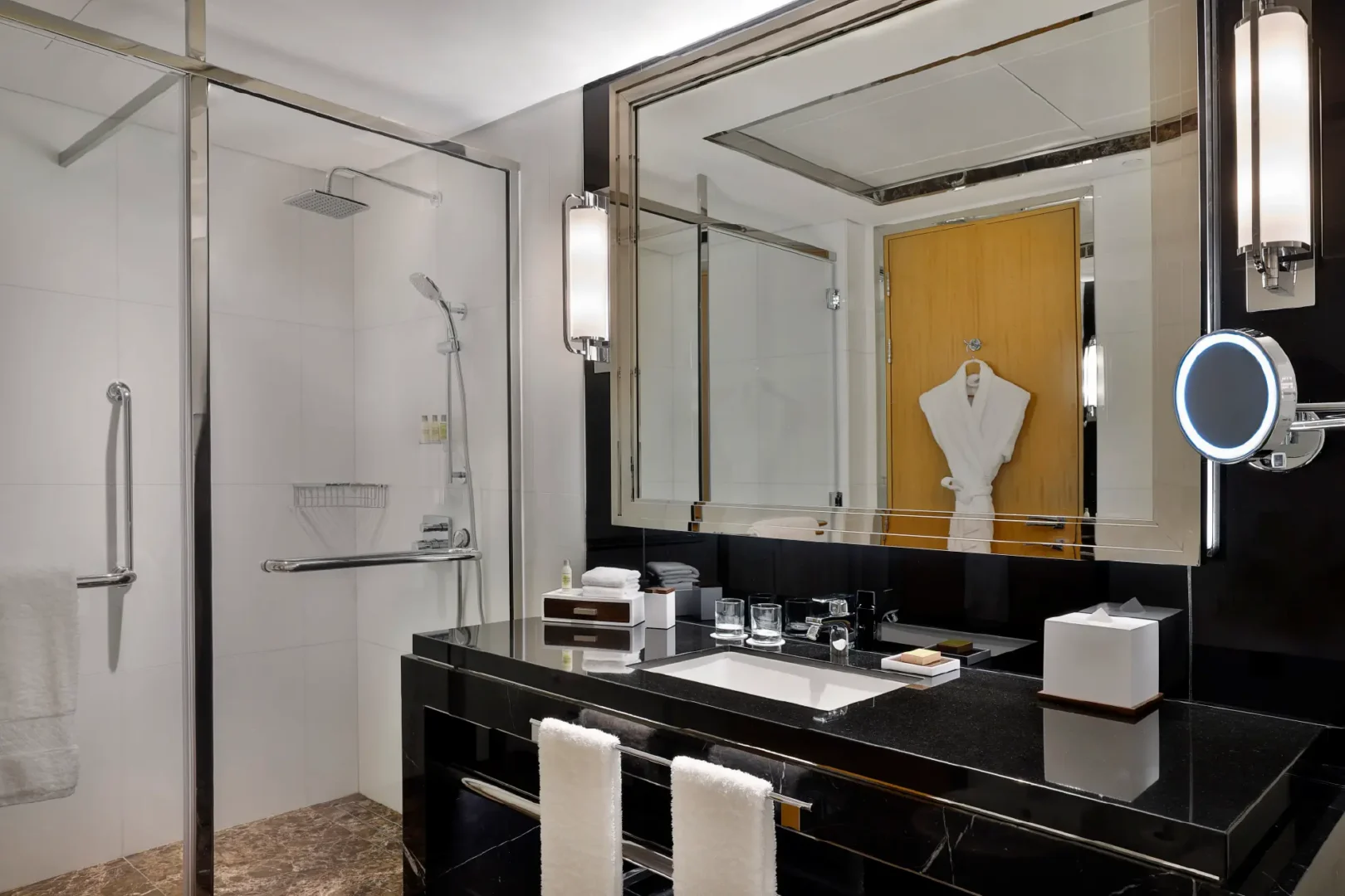 spacious hotel bathroom with walk in shower, black marble vanity, illuminated mirror and hanging bathrobe