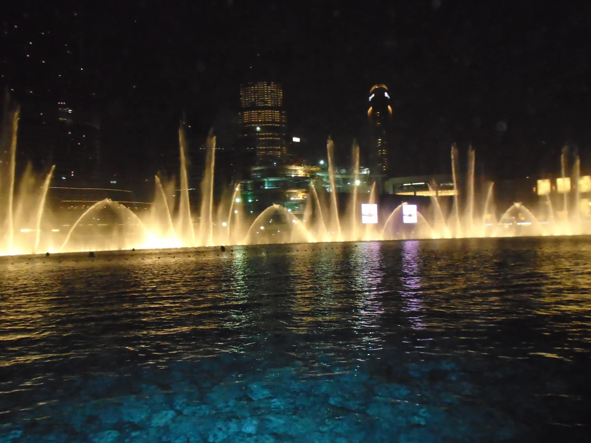 Dubai Fountain view from a small boat on the water at night with illuminated jets and city lights reflected on the surface